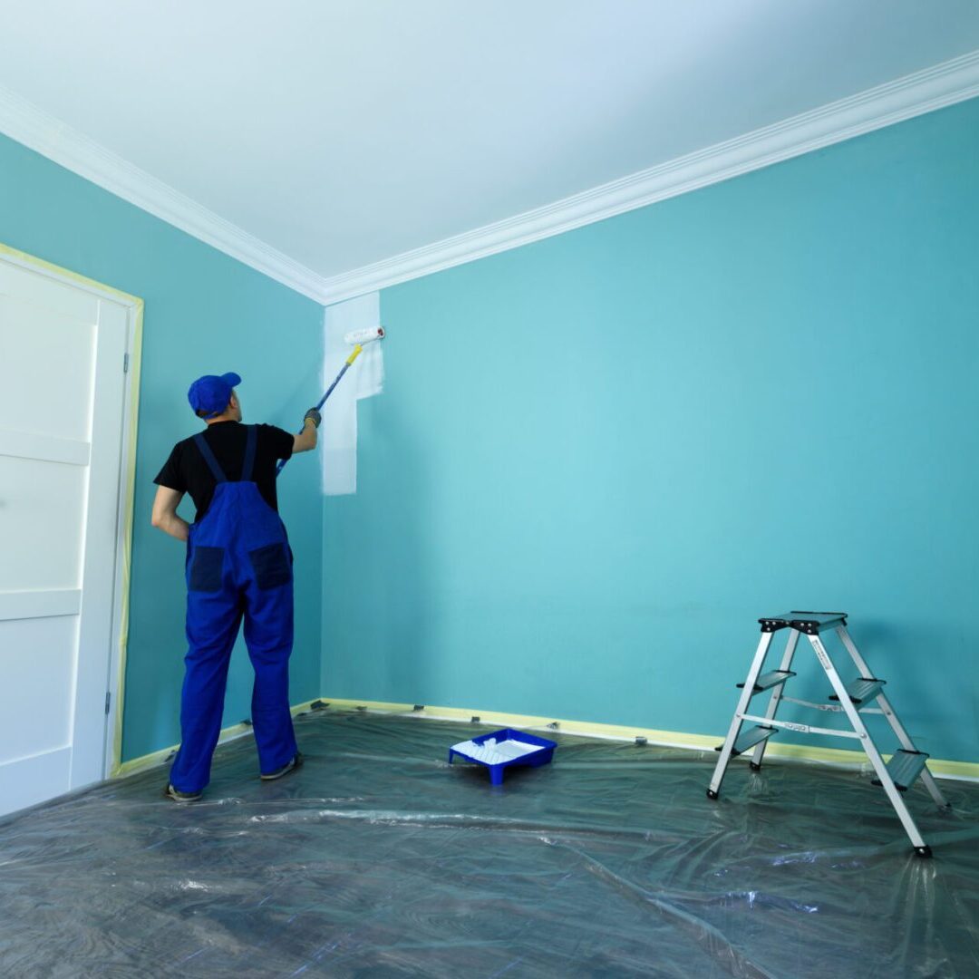 Man in a working overall Person painting a wall in blue room.